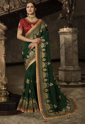 For A Proper Traditional Look, Grab This Attractive Looking Heavy Designer Saree In Dark Green Color Paired With Contrasting Red Colored Blouse. This Saree And Blouse are Silk Based Beautified With Heavy Jari And Resham Embroidery with Stone And Sequence Work. 